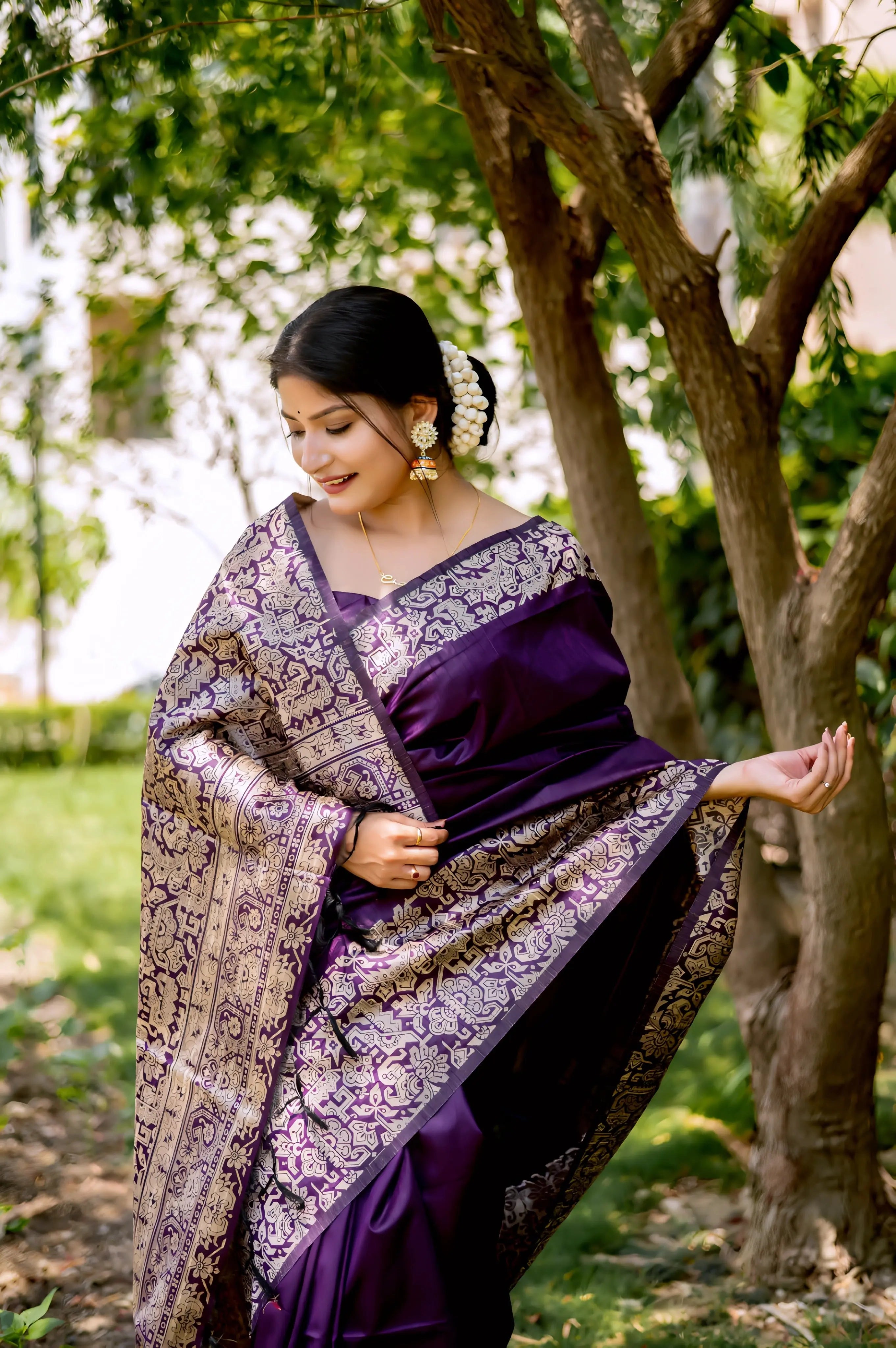 A woman models a luxurious Royal Purple Raw Silk Saree. The saree features a wide border and pallu with dense metallic (silver/light gold) Zari weaving in a Kalamkari-style floral and paisley pattern. She wears a matching purple short-sleeve blouse and gold jewelry, posing outdoors on a stone pathway.